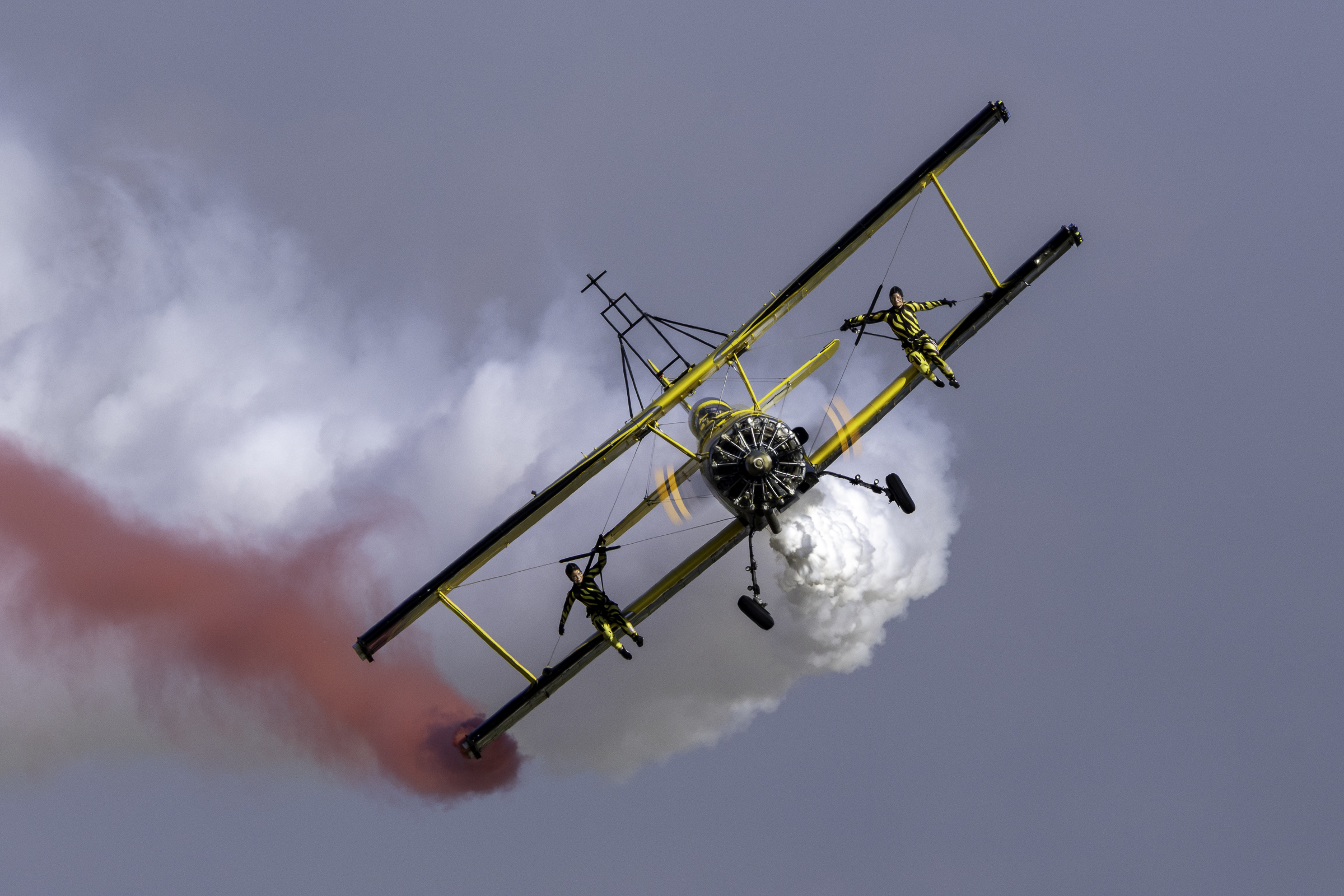 Two wingwalkers standing on the wings of a yellow-and-black biplane trailing red and white smoke