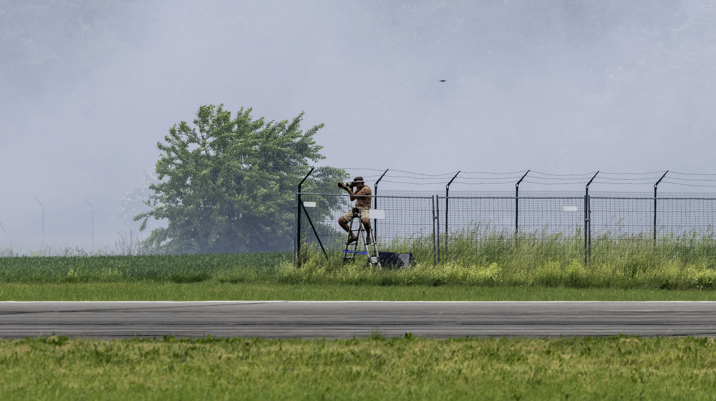 Photographer on a stepladder at the airfield fence shooting through drifting smoke